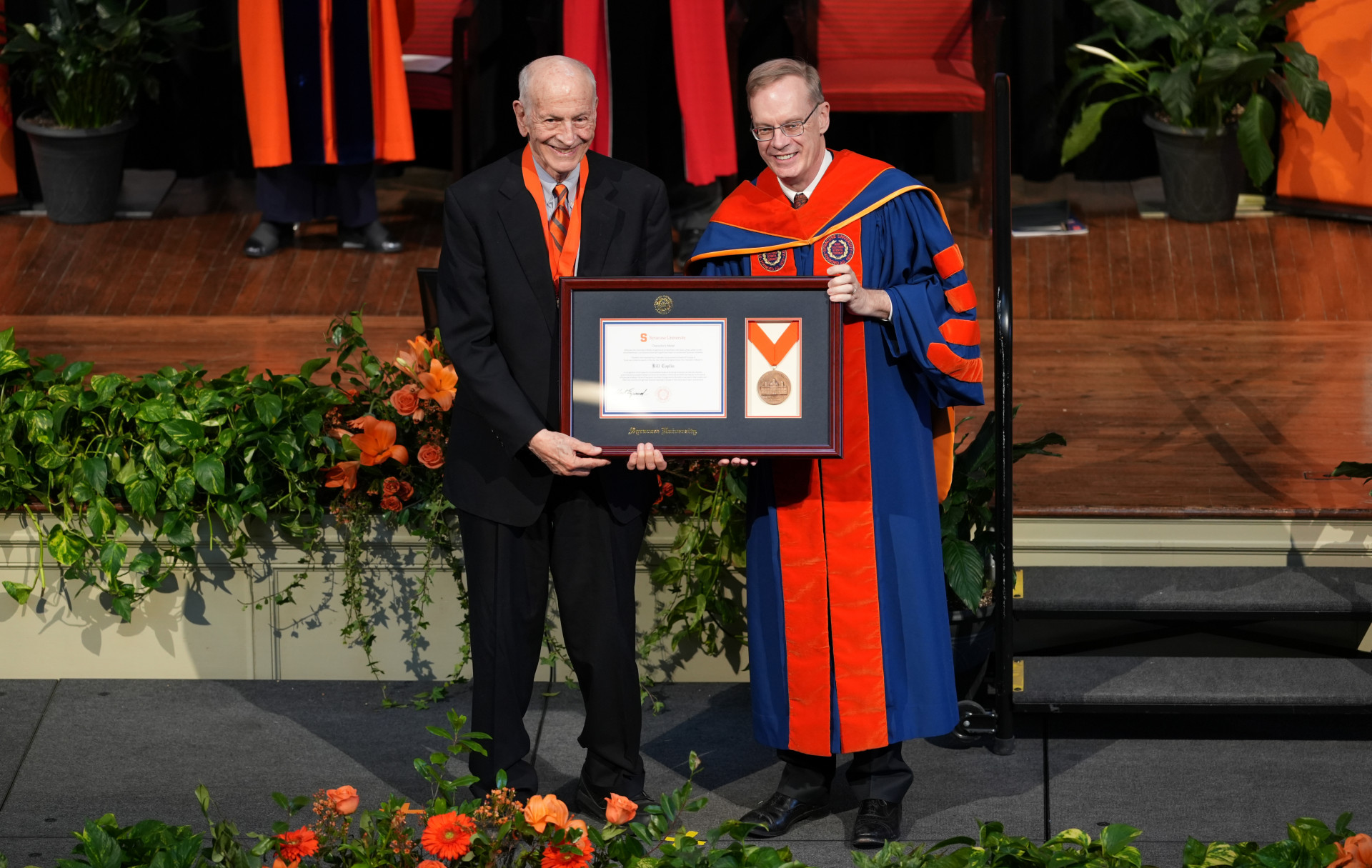Professor Bill Coplin and Chancellor Kent Syverud stand on a stage holding a framed certificate at the One University Awards. Syverud is wearing academic regalia, including a blue and orange gown with a hood, while Coplin is dressed in formal attire, including a black suit and tie. The stage is decorated with green plants and orange flowers.