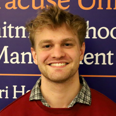 A person is standing in front of a blue background with orange text. The text reads: "Whitman School of Management" and "Syracuse University." The person is wearing a red sweater over a checkered shirt.