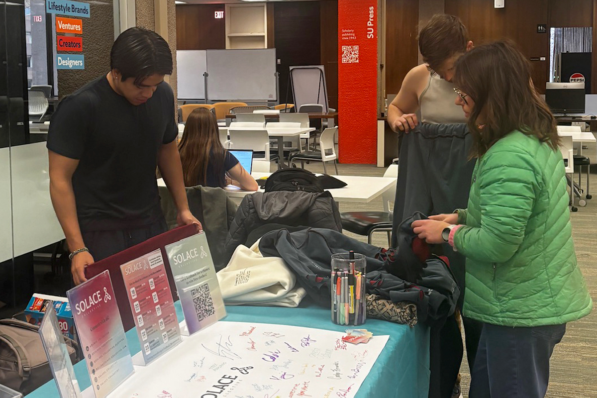The image shows three people standing around a table in what appears to be a communal workspace or library. The table is covered with various items, including informational posters, a large sheet of paper with signatures, and containers holding pens and markers. One person is looking at the posters on the table, another person is holding up a piece of clothing, and the third person is engaged in conversation while wearing a green jacket. In the background, there are more tables and chairs, as well as shelves with books or other materials. The setting looks like an organized event or workshop.